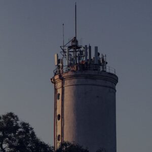 Close-up of an industrial tower with antenna installations at sunset, surrounded by trees.
