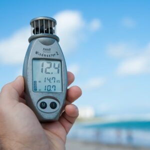 Close-up of a hand holding Windmaster 2 anemometer against a clear blue sky and beach backdrop.